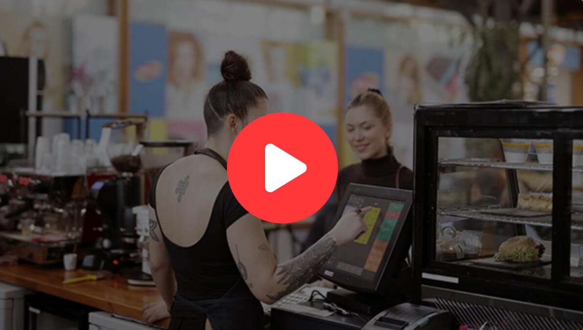 Woman with tattoos entering order into POS terminal with female customer smiling next to cake stand, orange play button over image