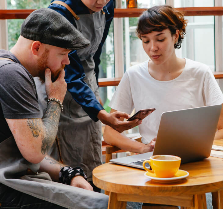 Waiter discussing mobile phone with other casually dressed employees, with laptop and coffee on small round table